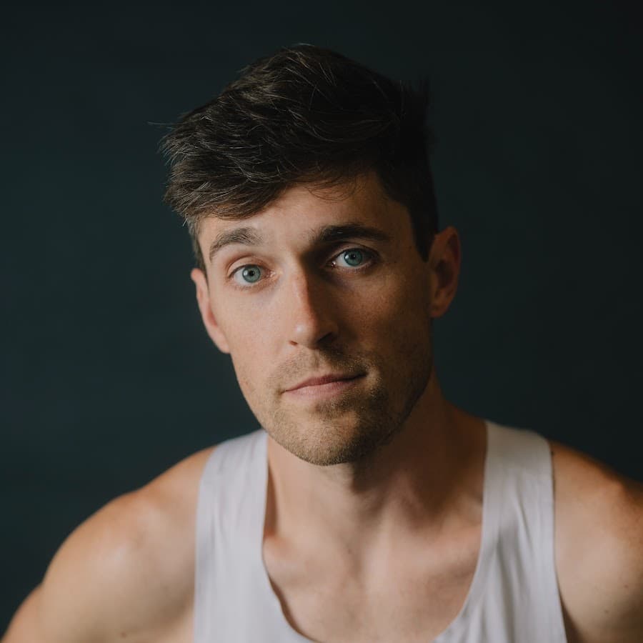 Close-up portrait of a man in a white singlet looking at camera against a dark neutral background, soft studio light on his face.