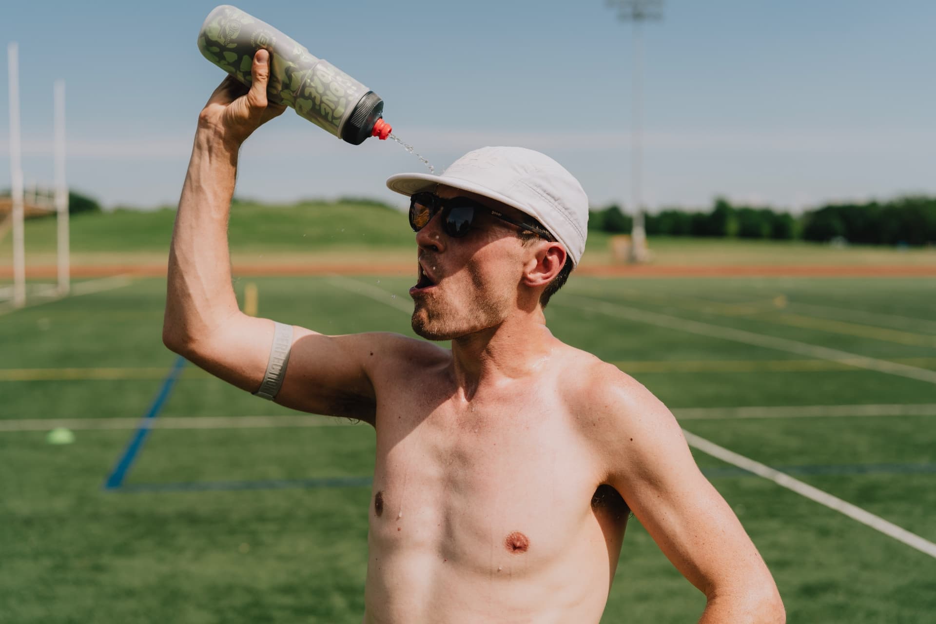 Shirtless runner in a white cap and sunglasses pours water from a bottle over his face on a green sports turf on a sunny day.