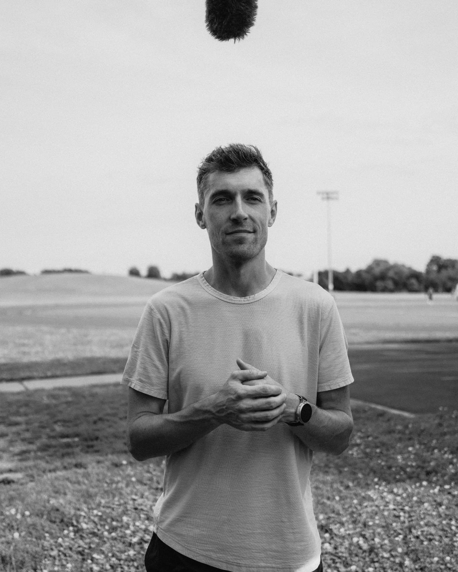 A man stands facing the camera on a grassy field with a boom microphone visible above him, overcast sky and sports lighting in the background.