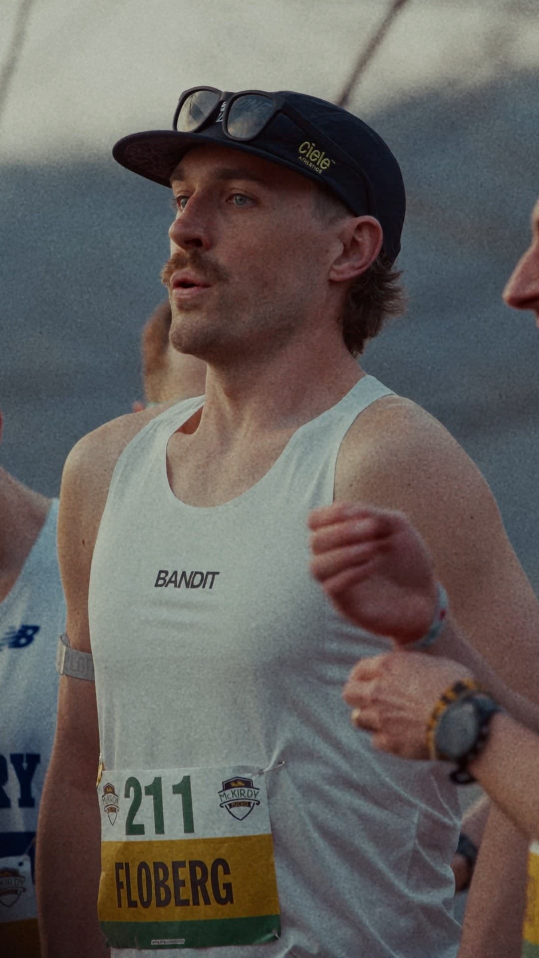 Male runner in a white singlet and black cap wearing bib 211 and sunglasses on his cap, standing among other runners in soft evening light.