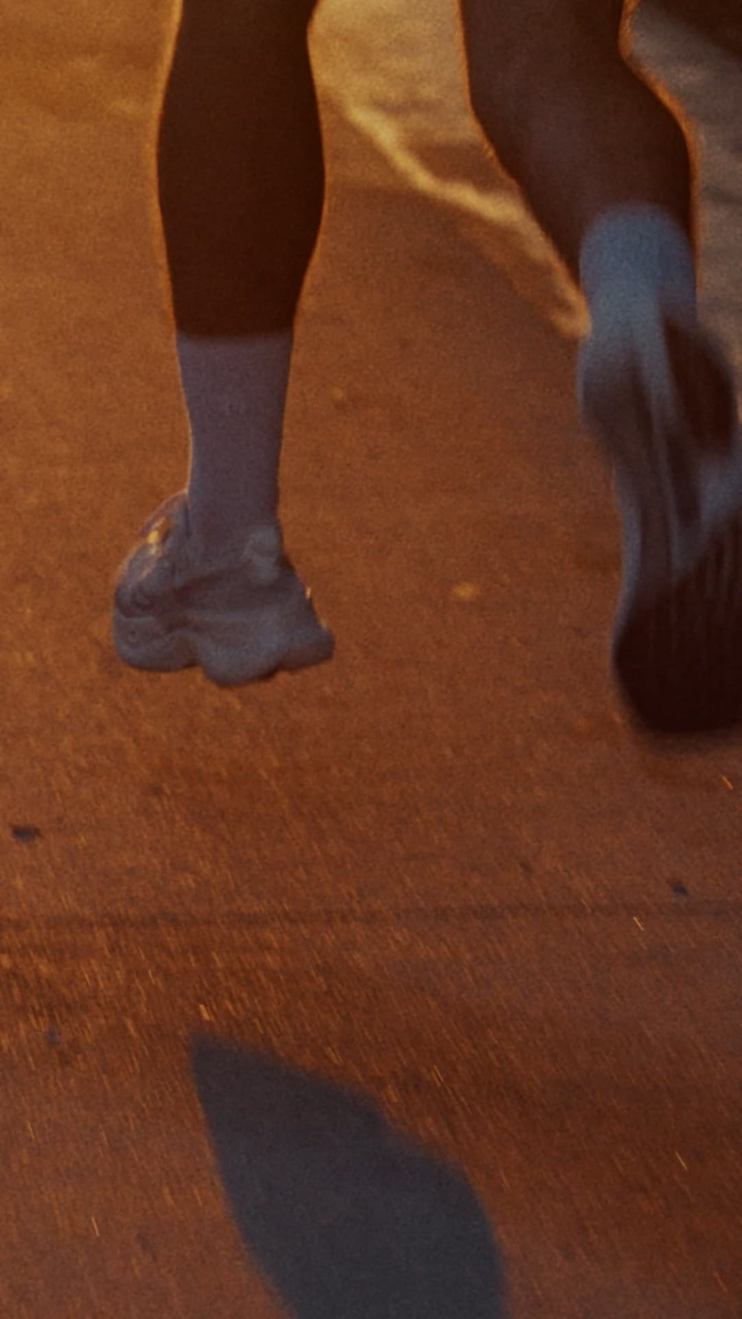 Low-angle close-up of two runners' lower legs and shoes mid-stride on a warmly lit pavement, ankle shadow visible on the ground.