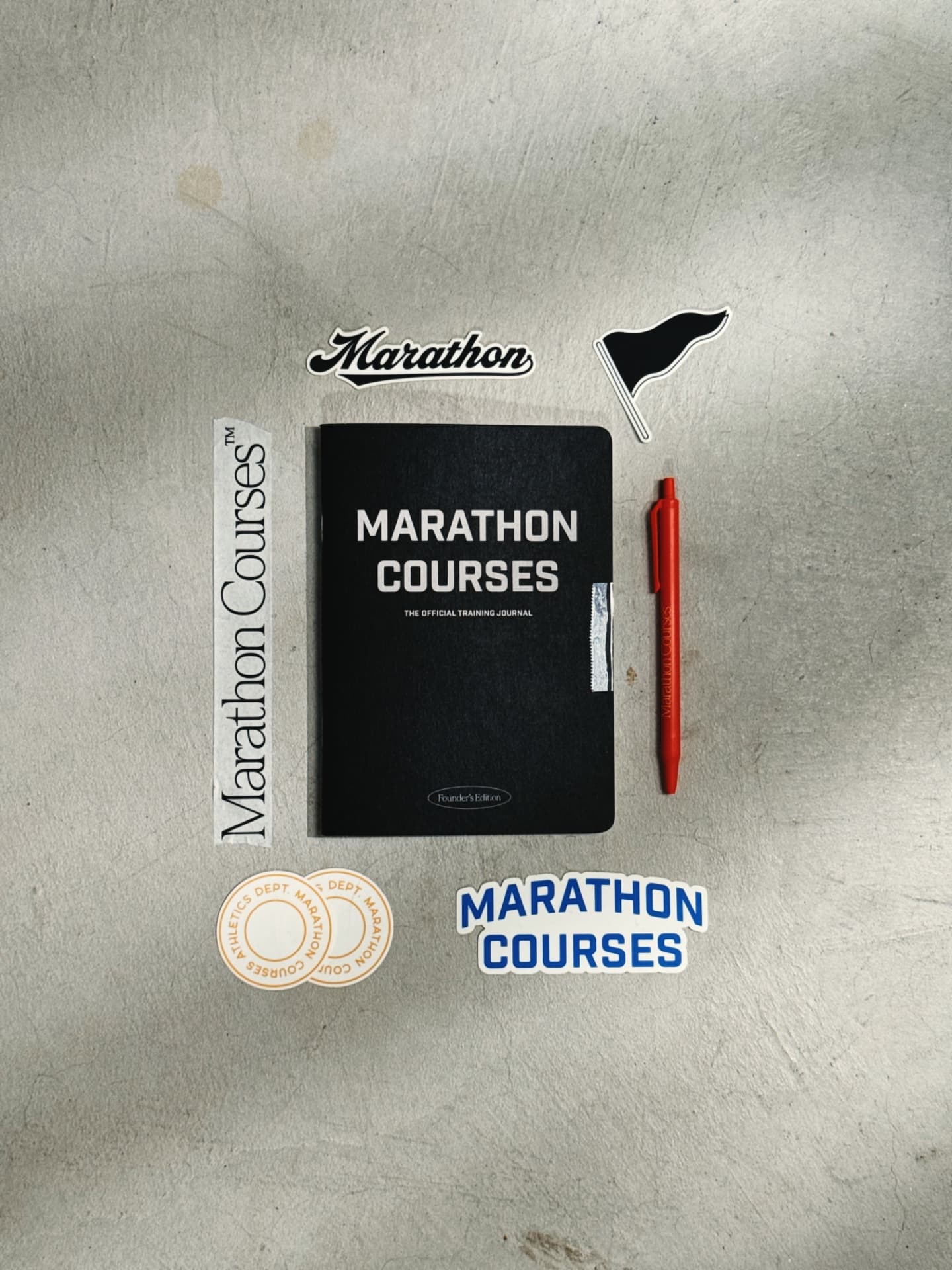 Flat-lay of a black 'Marathon Courses' training journal surrounded by brand stickers and a red pen on a grey concrete surface.